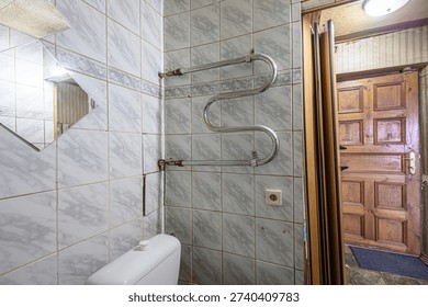 A small tiled bathroom with a marble-patterned wall, curved water полотенцесушитель, toilet, mirror, and an open door leading to a room with a wooden door. Clean, functional layout