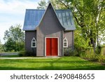 A small or tiny house with grey exterior wooden walls, small clear glass church windows, metal peaked roofs and vibrant red double doors. The building