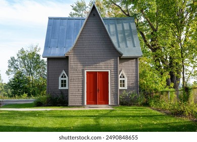 A small or tiny house with grey exterior wooden walls, small clear glass church windows, metal peaked roofs and vibrant red double doors. The building's garden is green grass and tall lush trees. - Powered by Shutterstock