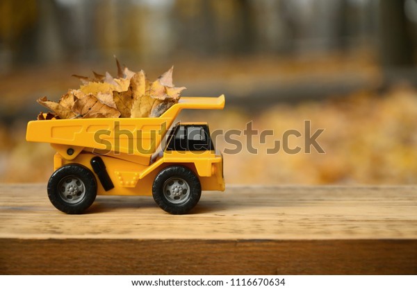 A small toy yellow truck is loaded with yellow
fallen leaves. The car stands on a wooden surface against a
background of a blurry autumn park. Cleaning and removal of fallen
leaves. Seasonal works