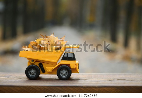 A small toy yellow truck is loaded with yellow\
fallen leaves. The car stands on a wooden surface against a\
background of a blurry autumn park. Cleaning and removal of fallen\
leaves. Seasonal works