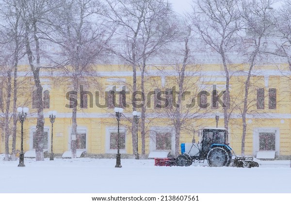 A small tractor with a snow plow removes snow\
in motion. Reduced visibility in a snowstorm. Old city. The\
building is yellow in the background. Street lamps in the old\
style. Urban winter\
landscape.