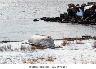 A small traditional fishing boat made of wood with white paint peeling. The worn vessel is overturned on an empty beach. The ground is covered in white snow. There's a rocky shoreline and beach.  - Powered by Shutterstock