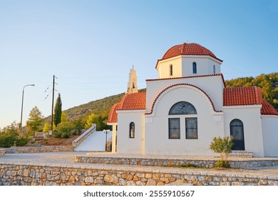 A small, traditional Greek Orthodox church with a red-tiled dome and white walls, set against a backdrop of green hills and a clear blue sky. - Powered by Shutterstock