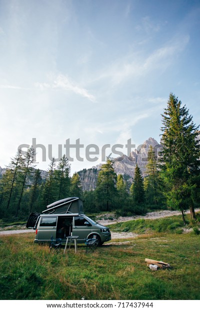 Small travel vehicle\
camping van or big car with folding rooftop with bed is parked on\
secluded wild site under huge mountain formation in dolomites,\
surrounded by forest