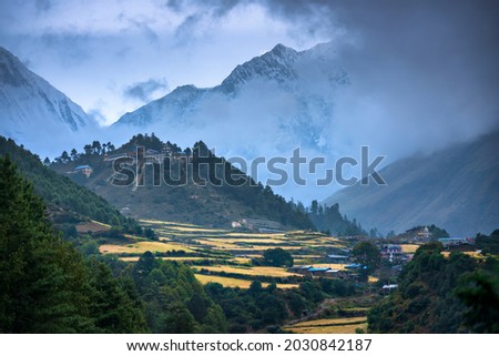 Small village on the mountain peak in fog at sunset in Nepal. Colorful landscape with houses, blue sky with low clouds, high rocks, green forest and yellow fields at dusk. Travel in Himalayas. Nature