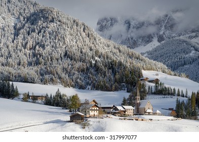 The small village of St. Magdalena with its church covered in snow and with the Dolomites mountains covered in the clouds in the Val di Funes Valley (Villnoesstal) in South Tyrol in Italy in winter.