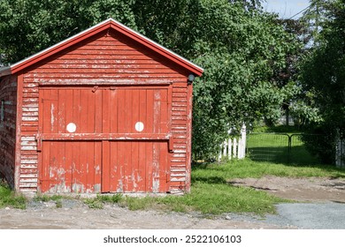 A small vintage red wooden garage. There are two exterior wood doors with white round circles painted on the building. The paint is worn and weathered on the structure. There are trees in the distance - Powered by Shutterstock