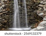 A small waterfall cascades down a wall of weathered rocks, moss adding a vibrant green hue. Water rushes over the stones, creating a soothing sound. The image captures the tranquility of nature