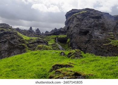 Small waterfall flowing from dark cliffs over a bright green grassy field in Iceland, under a cloudy sky