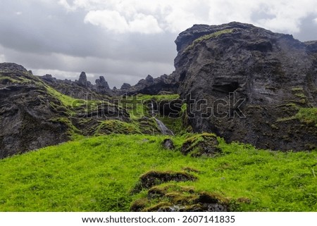Small waterfall flowing from dark cliffs over a bright green grassy field in Iceland, under a cloudy sky