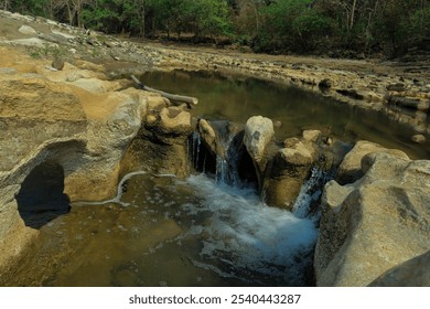 A small waterfall flows over uniquely shaped rocks in a shallow stream, creating a peaceful, scenic natural scene. The rocky formations and clear water provide a tranquil escape surrounded by nature - Powered by Shutterstock