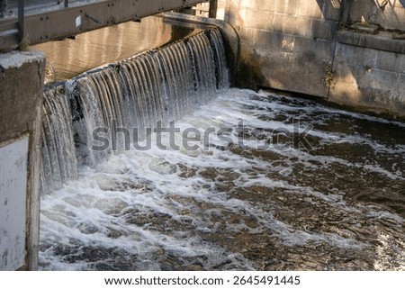 A small waterfall flows over a weir into a river. The structure demonstrates the combination of industrial construction and flowing water.
