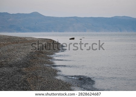 Small wavelets lap a gently curving pebble spit. Across the bay, hazy mountains emerge through the cool blue light of dawn.