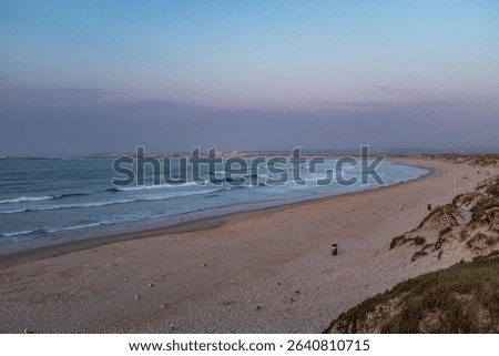 Small waves break gently on the wide beach of Praia da Baías, Portugal. In the background one can see the town of Ferrel, part of the municipality of Peniche, and the island of Baleal.