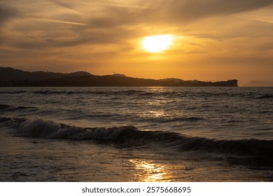 Small waves of the Ionic sea. Beach with pebbles. Mountain in the background. Sunset and cloudy sky. Reflection on the sea. Acharavi, Korfu, Greece.