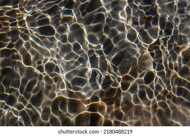 The small waves on the surface of the mountain stream bend the sun's rays. This creates bright light lines. In these patterns, the light is refracted and rainbows are created over the sandy streambed.