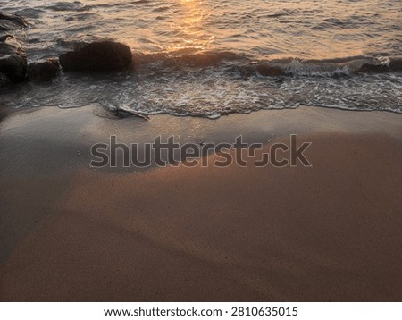 Small waves rolling onto a sandy shoreline with coastal rocks illuminated by the warm glow of the setting sun.