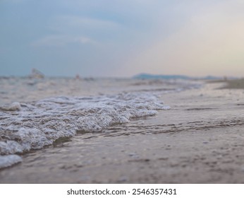 Small waves rolling through shallow water and creating foam. The water is clear. Coastline or mountains in the distance. The sky is painted in soft pastel colors of blue and light yellow.