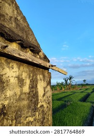 a small, weathered brick structure, likely an old hut or storage shed, standing beside an expansive agricultural field under a clear blue sky. The building features exposed, reddish-brown bricks on its side wall, while the front triangular gable is covered in a distressed, gray plaster coating that shows signs of aging and discoloration.