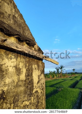 a small, weathered brick structure, likely an old hut or storage shed, standing beside an expansive agricultural field under a clear blue sky. The building features exposed, reddish-brown bricks on its side wall, while the front triangular gable is covered in a distressed, gray plaster coating that shows signs of aging and discoloration.
