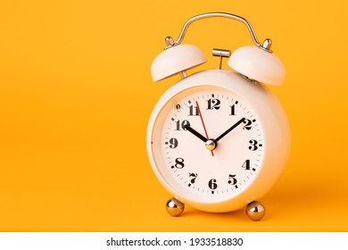 Small white alarm clock, black numbers, set the time placed on a table. Clock on isolated yellow background.