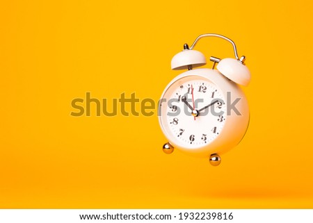 Small white alarm clock, black numbers, set the time placed on a table. Clock on isolated yellow background.