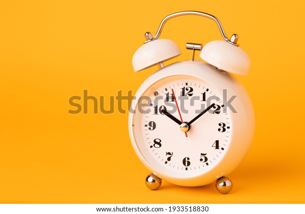 Small white alarm\
clock, black numbers, set the time placed on a table. Clock on\
isolated yellow\
background.
