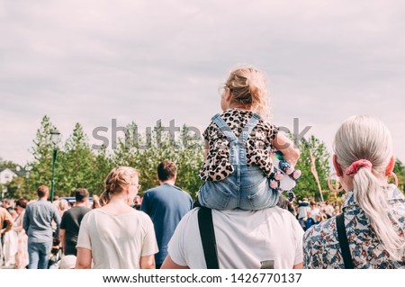 Small white blond child girl kid sitting on the fathers parent shoulders in the summer day with people walking around and green trees on the background