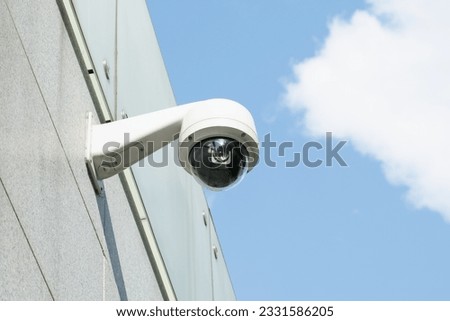 Small white CCTV camera camera on the wall of building against a blue sky . Security, video surveillance, face recognition. Providing security in a residential area.
