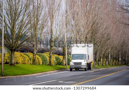 Small white compact useful transportation commercial light-duty local haul rig truck with box trailer transporting local goods on the city road with tree alley and railroad