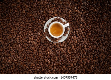 Small white cup of frothy hot strong espresso coffee nestling on a layer of full roasted coffee beans in a full frame background texture - Shutterstock ID 1407559478