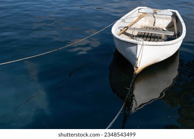Small white fishing boat tied with a rope, floats in dark blue water. . Boat mirrored in water. Space for text or design