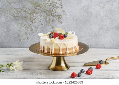 Small White And Milk Chocolate Mousse Cake Decorated With Fresh Berries On Golden Cake Stand Over Gray Background With Flowers And Berries. Side View, Copy Space