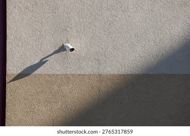 Small white security camera mounted on a textured two-tone wall casting a long shadow. Minimalist urban surveillance and safety concept under bright sunlight.