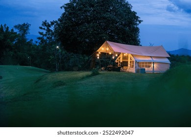 A small white tent is set up in a grassy field. The tent is lit up with lights, giving it a cozy and inviting atmosphere. The scene is peaceful and serene, with the tent providing a sense of shelter