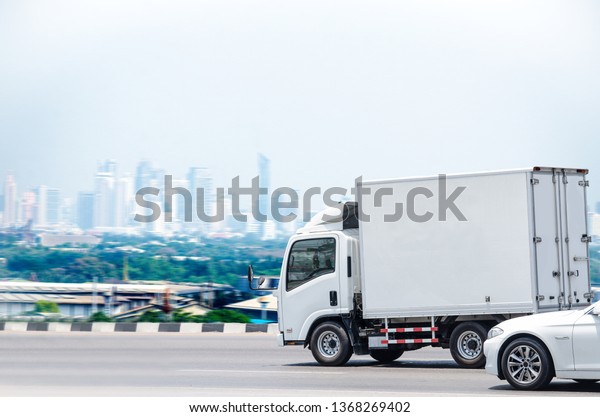 A small white truck running on\
the road for a transportation business with a city\
background.