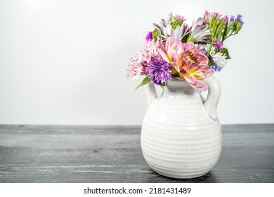 Small White Two-handled Jug Vase Filled With A Variety Of Home Grown Flowers. White Background And Weather Black Wood Base.