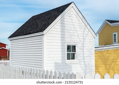 A small white wooden shed with a single window in the center and black shingles on the roof in a garden with a white picket fence. There are yellow and red buildings and blue sky in the background.   