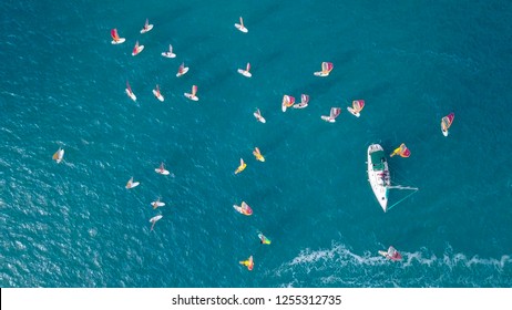 Small White Yacht Surrounded By A Large Group Of Wind Surfers - Top Down Aerial Image.