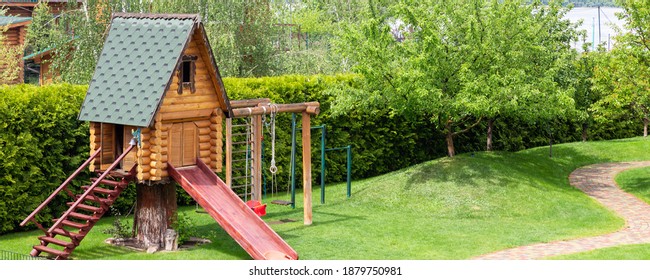 Small wood log playhouse hut with stairs ladder and wooden slide on children playground at park or house yard. Panoramic view. Green grass lawn garden and paved pathway background on bright sunny day