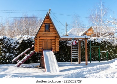 Small Wood Log Playhouse Teehouse Hut Snowcapped Stairs Ladder Wooden Slide Children Playground At Park Or House Yard. White Snow Covered Lodge Blue Clear Sky Background On Bright Sunny Winter Day