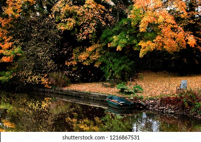 Small Wooden Boat In The River Ollen, Germany Surrounded By Big Trees In Beautiful Colored Autumn Foliage, A Garden Chair Is Standing Next To The Water, Too