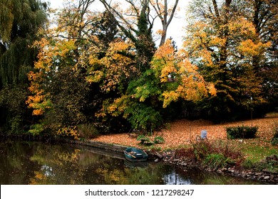 Small Wooden Boat In The River Ollen, Germany Surrounded By Big Trees In Beautiful Colored Autumn Foliage, A Garden Chair Is Standing Next To The Water, Too