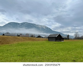 A small wooden cottage rests on a meadow, framed by the Alps mountain range in the rural Partnachklamm Gorge area of Garmisch-Partenkirchen, Germany, under a cloudy sky. - Powered by Shutterstock