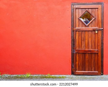 Small Wooden Door With Square Window And Lock And Red Color Wall. Rich Saturated Colors. Copy Space. Backdoor To A Garden Or Small Shed.