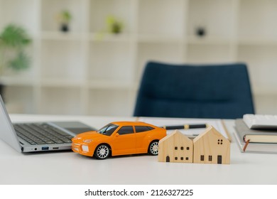 Small Wooden House On The Table And Orange Car And Laptop. Blurry Background In The Office Real Estate Industry Leasing Concept