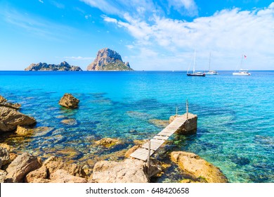 Small wooden pier in Cala d'Hort bay and view of Es Vedra island, Ibiza island, Spain