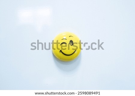 A small yellow smiley face button with a winking expression on a white background. The minimalistic composition highlights the playful and cheerful nature of the emoji-inspired design