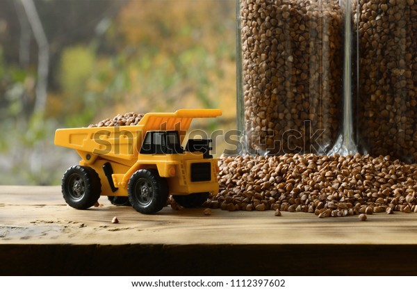 A small yellow toy truck is loaded with brown\
grains of buckwheat around the buckwheat pile and a glass of croup.\
A car on a wooden surface against a background of autumn forest.\
Buckwheat delivery